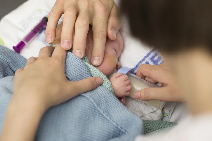 Spedbarn i seng som får flaske og omsluttes av hender